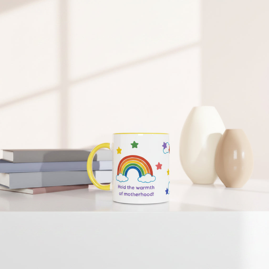 A white ceramic mug with a yellow handle and rim, decorated with a colorful rainbow, stars, clouds, and the text 'Hold the warmth of motherhood!', styled on a white table next to books and two ceramic vases.