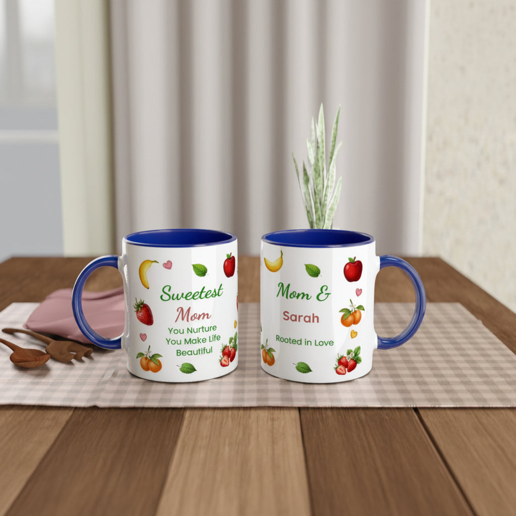 Two personalized ceramic mugs with blue interiors and handles, displayed on a wooden table with a placemat and small decorative props. The mugs feature fruit illustrations and custom names and messages: one reads 'Sweetest Mom You Nurture You Make Life Beautiful' and the other reads 'Mom & Sarah Rooted in Love'.