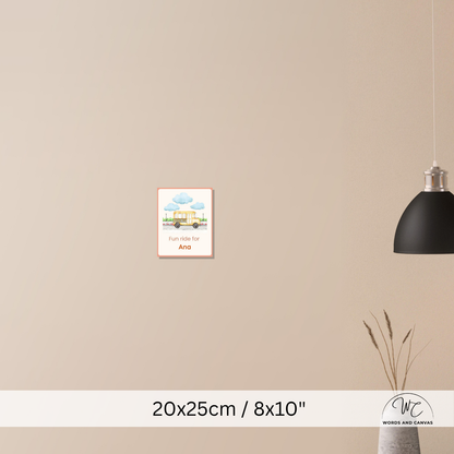 A small framed wall art print hangs on a beige wall in a styled interior. The print shows a colourful illustration of a house under blue clouds with the text 'Fun ride for Ana'.