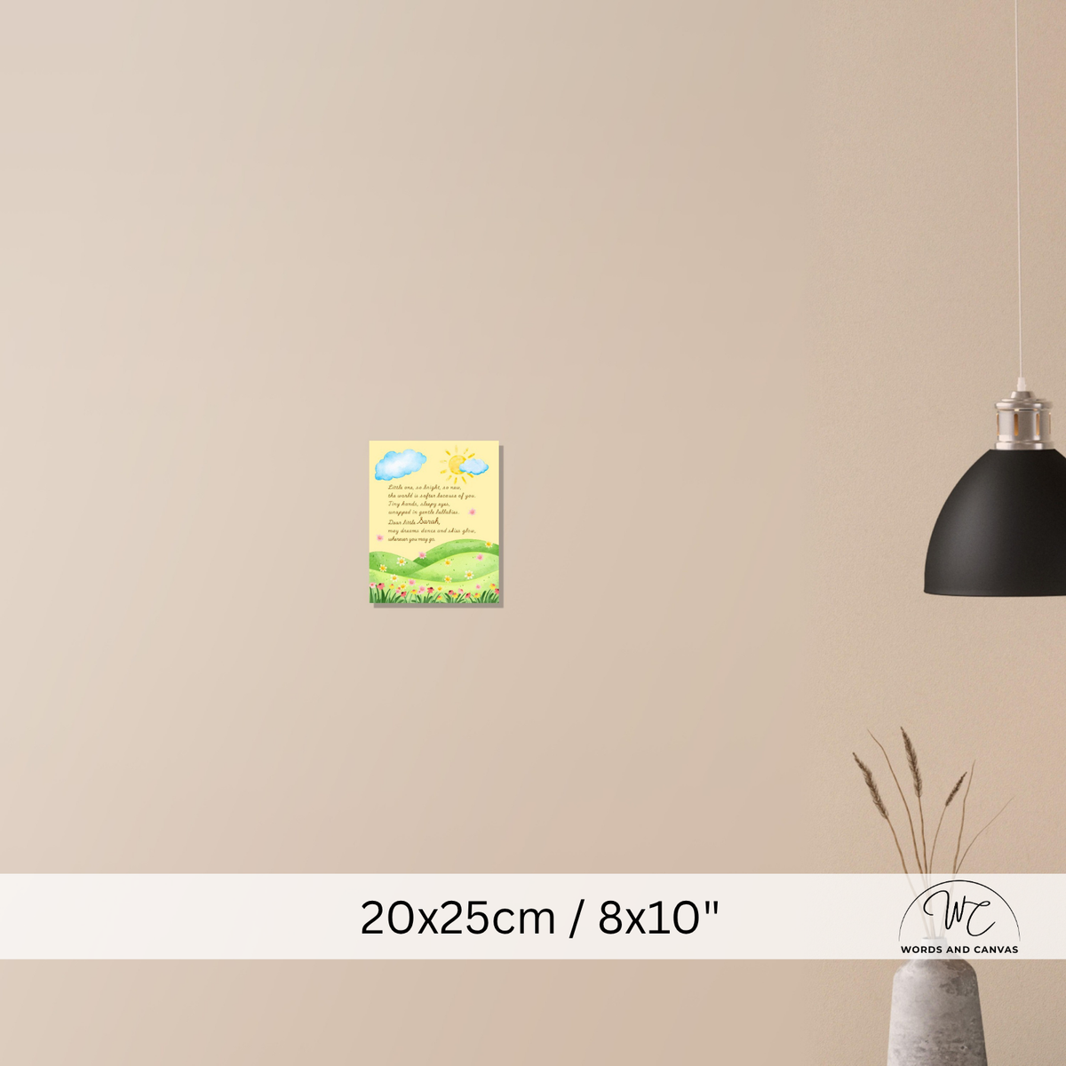 A small vertical wall art print with a yellow sky, blue clouds, green hills, and cursive text, shown mounted on a beige wall in a styled interior. A black pendant light hangs on the right, and a vase with dried stems sits below.