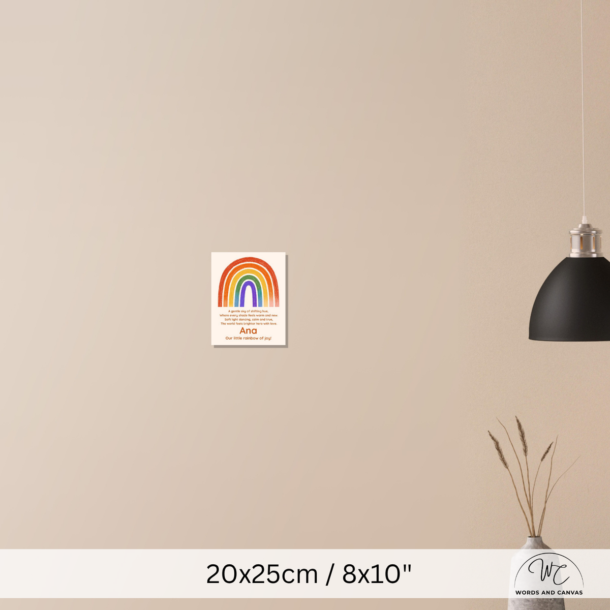 A small vertical rainbow art print with text is mounted on a beige wall in a styled interior scene. A black pendant lamp hangs on the right, and a vase with dried stems sits near the bottom right.