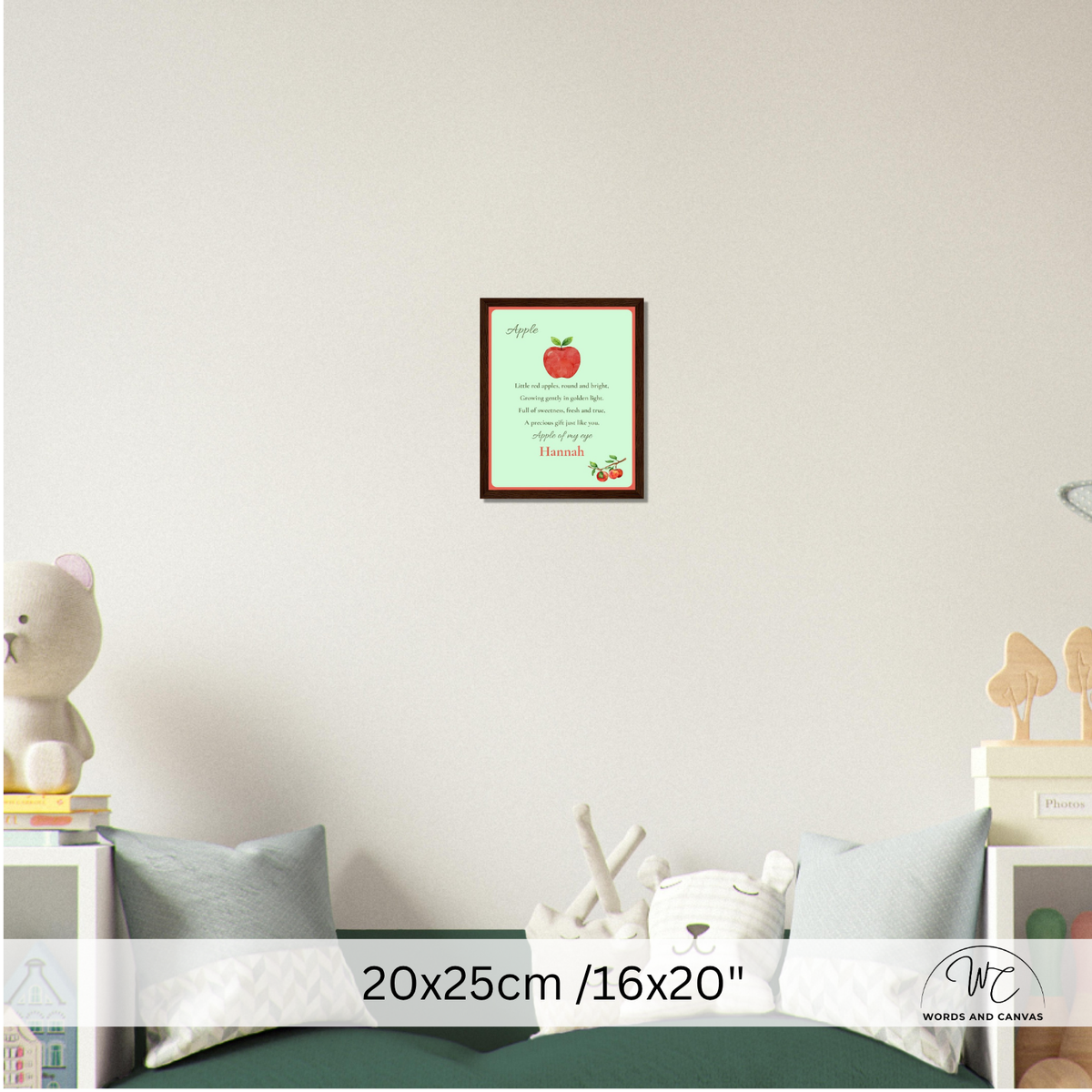 A framed apple-themed nursery wall art print hanging on a white wall above a bed. The print features a red apple with the word 'apple' and a poem with the name 'Hannah' at the bottom.