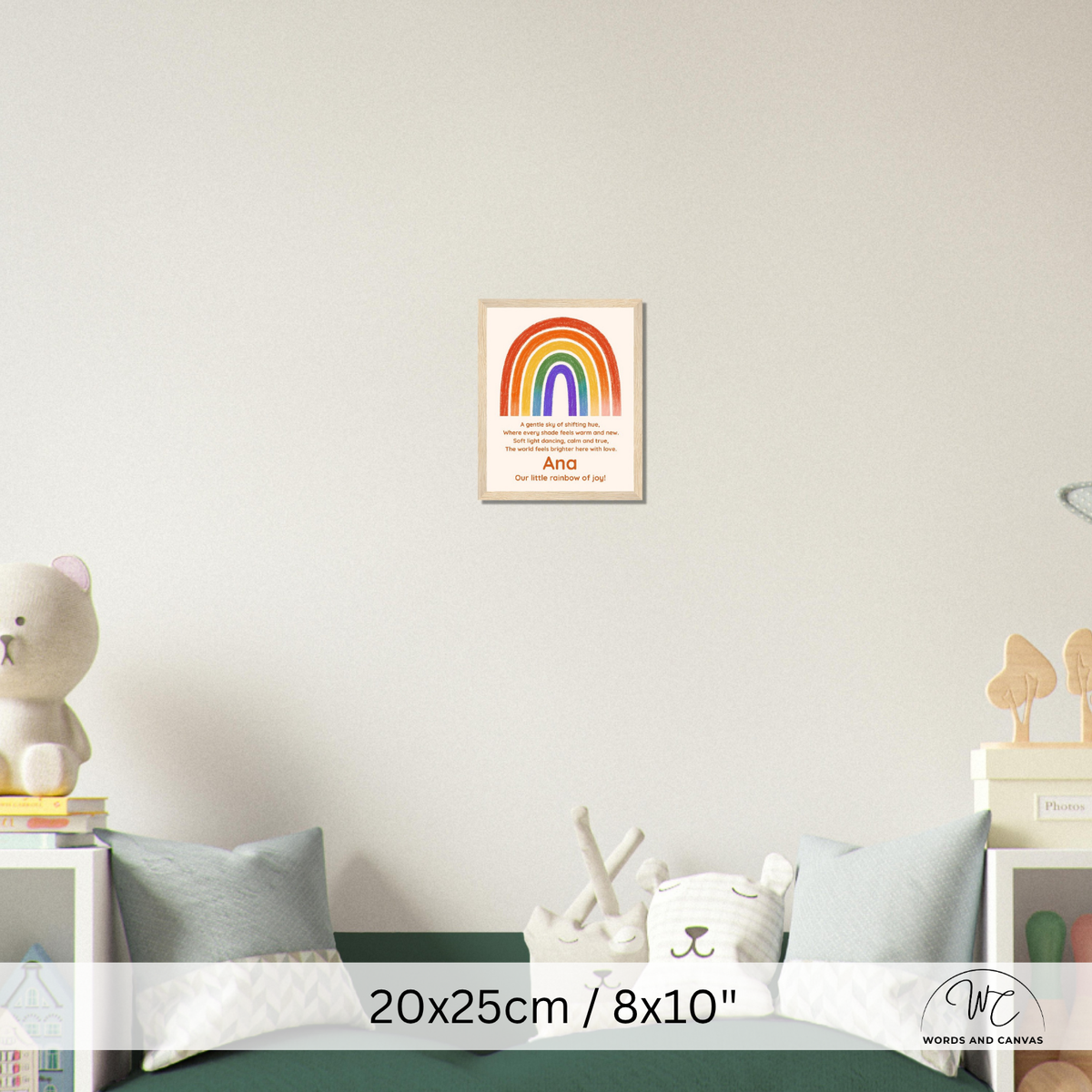 A framed children's wall poster featuring a colorful rainbow and the word 'kind' displayed in a nursery setting above a bed. The poster is shown in a lifestyle context with a teddy bear, pillows, and bedside tables.