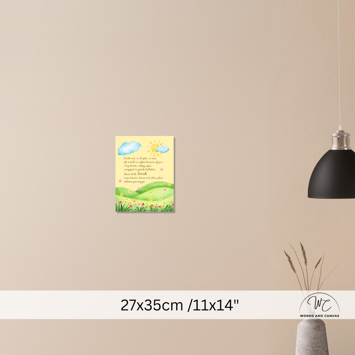 A small wall art print with a yellow background, blue clouds, green hills, and handwritten-style text is shown hanging on a beige wall. A black pendant lamp hangs to the right, and a vase with dried stems sits below.
