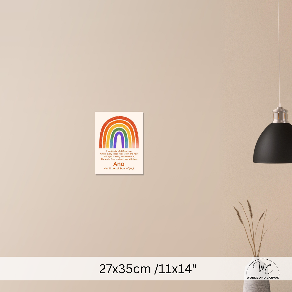 A personalized rainbow wall art print with a poem, displayed on a beige wall in a styled room setting with a black pendant lamp and a vase with dried grass. The print features a colorful rainbow and the name 'Ana'.