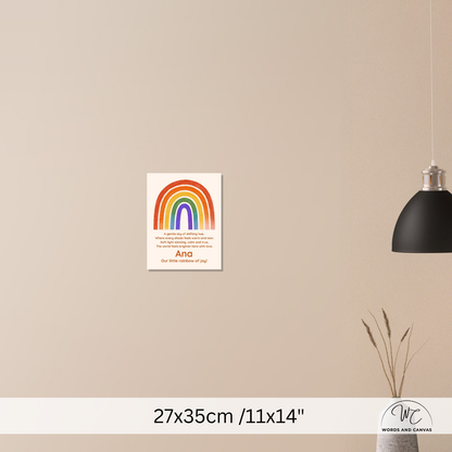 A personalized rainbow wall art print with a poem, displayed on a beige wall in a styled room setting with a black pendant lamp and a vase with dried grass. The print features a colorful rainbow and the name 'Ana'.