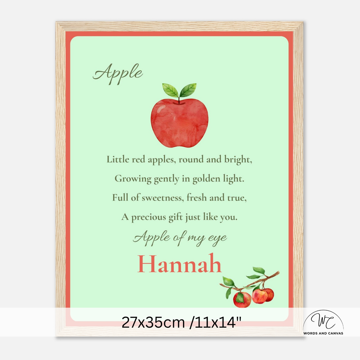 A personalized framed apple-themed nursery print featuring a watercolor red apple, nursery rhyme text, and the name 'Hannah' in coral red, shown with a light wood frame on a white background.