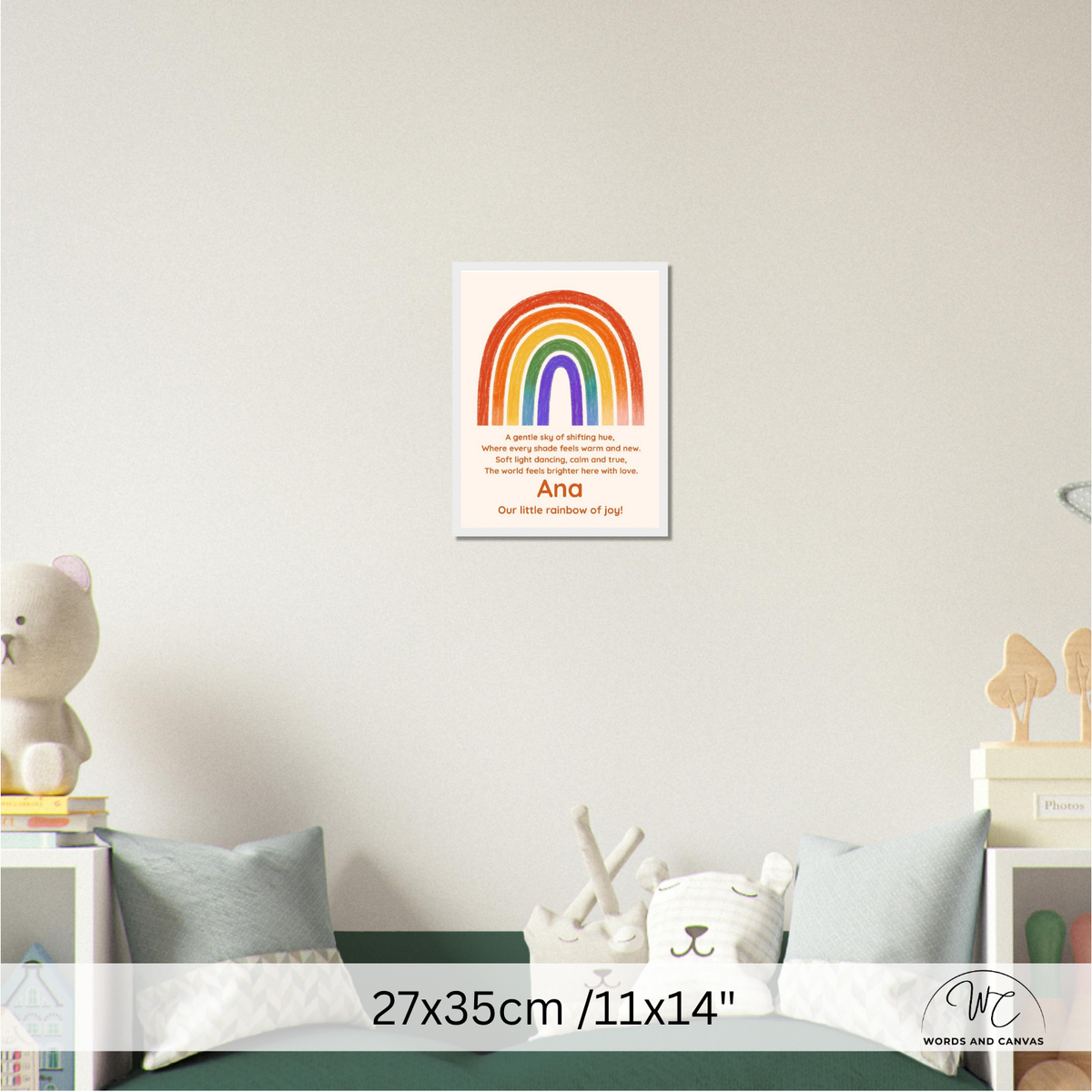 A personalized rainbow wall art print displayed on a light gray wall above a child's bed. The poster features a colorful rainbow arch with the text 'Our little rainbow of joy' and the name 'Ana'.