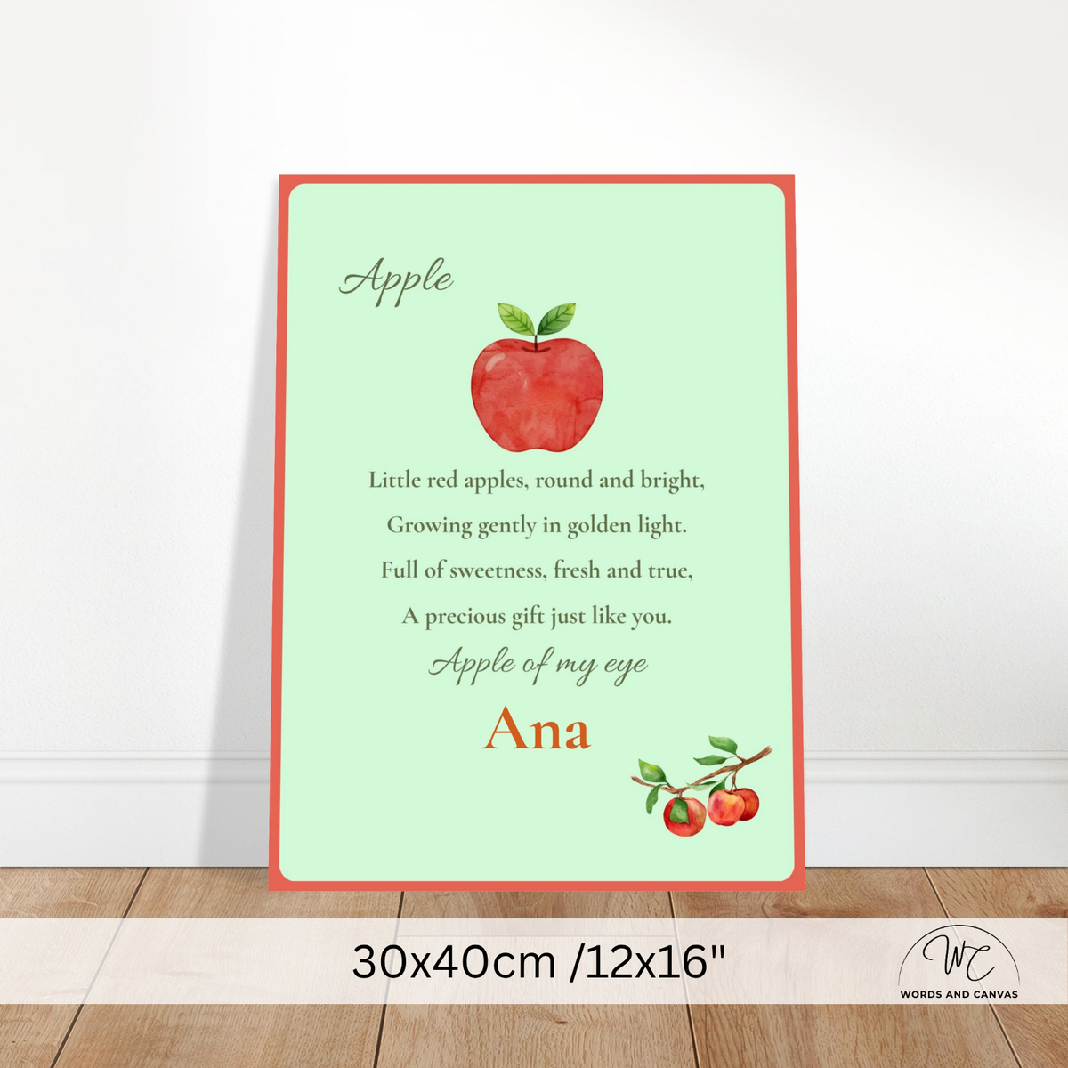 A personalized apple-themed nursery print with a red border, a central red apple illustration, and a poem ending with the name 'Ana'. The print is shown leaning against a white wall on a wooden floor, with a size overlay at the bottom indicating 30x40cm /12x16".