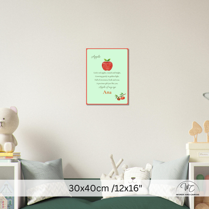 A personalized apple-themed nursery wall art print displayed on a light grey wall above a bed. The print features a light green background with a red border, a red apple illustration, and the name 'Ana' 