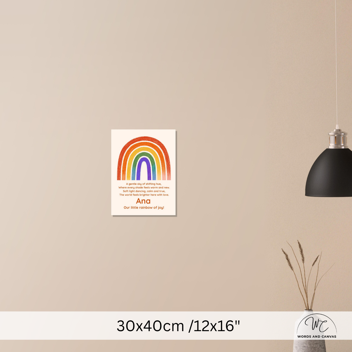 A personalized rainbow wall art print with a poem, displayed on a beige wall in a styled room setting with a black pendant lamp and a vase with dried grass. The print features a colorful rainbow and the name 'Ana'.