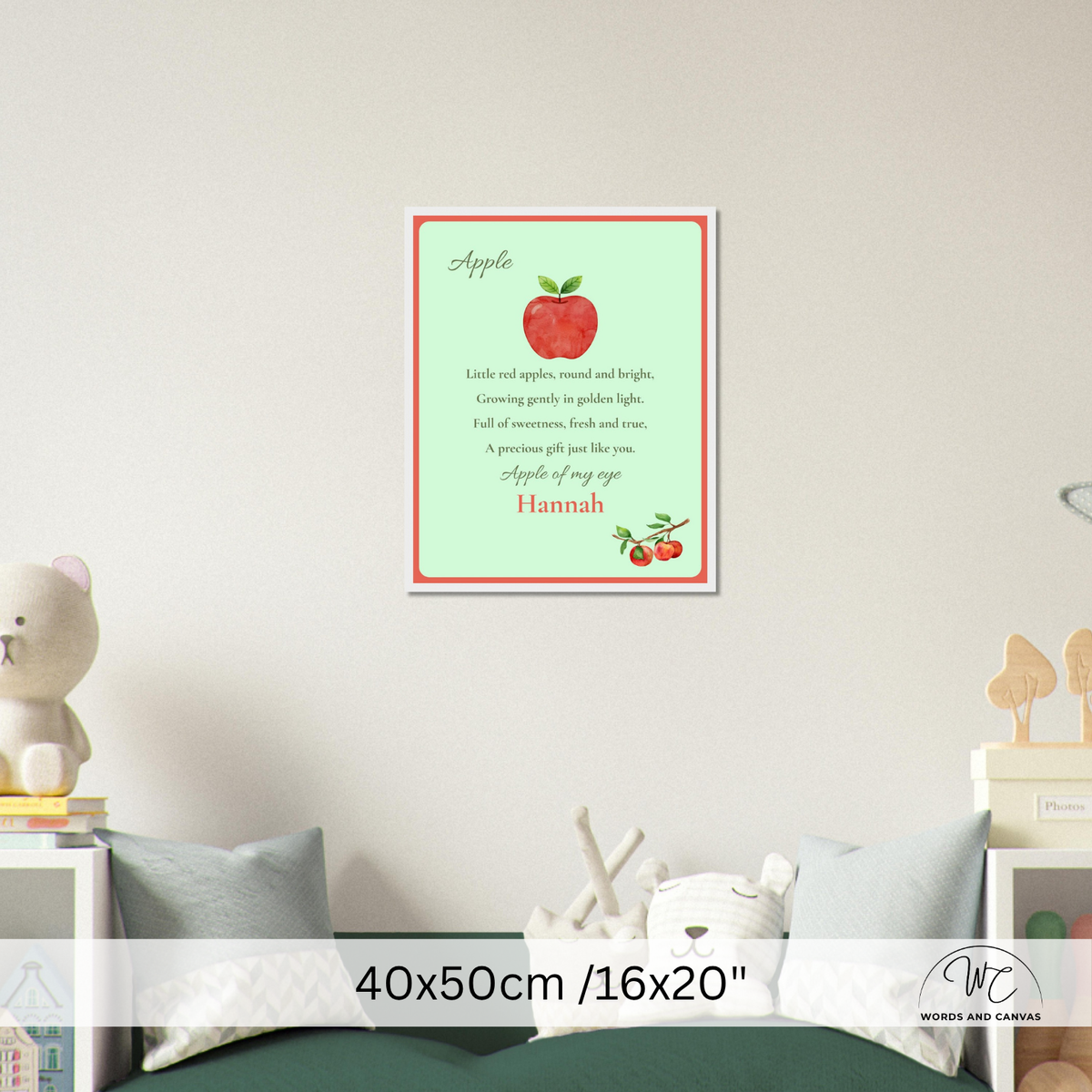 A framed personalized apple-themed nursery wall art print hanging on a light beige wall above a bed. The print features a red apple and cherries on a pale green background with a red border, and the text 'Apple' and a custom name, 'Hannah'.