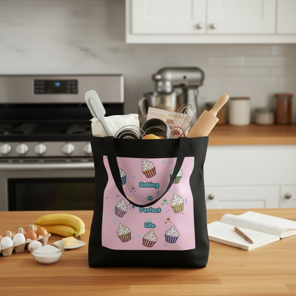 A black tote bag with a pink cupcake print panel sits on a wooden kitchen counter, filled with baking utensils. The kitchen background is softly blurred, showing white cabinets, a stove, and a stand mixer.
