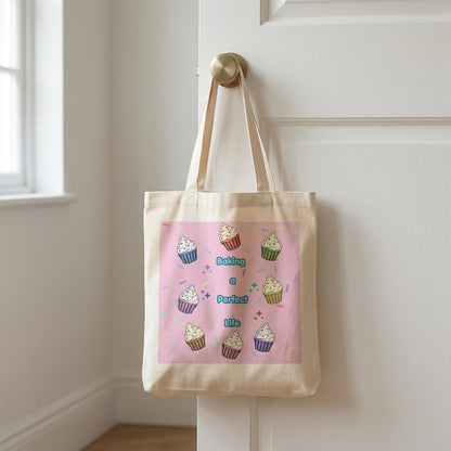 A canvas tote bag with a light pink printed panel featuring illustrated cupcakes and the words 'Baking a Perfect Life' in blue, hanging from a brass doorknob on a white paneled door.