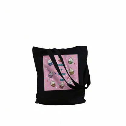 A black canvas tote bag with a square pink graphic print on the front featuring illustrated cupcakes and the text "Baking a perfect Life"