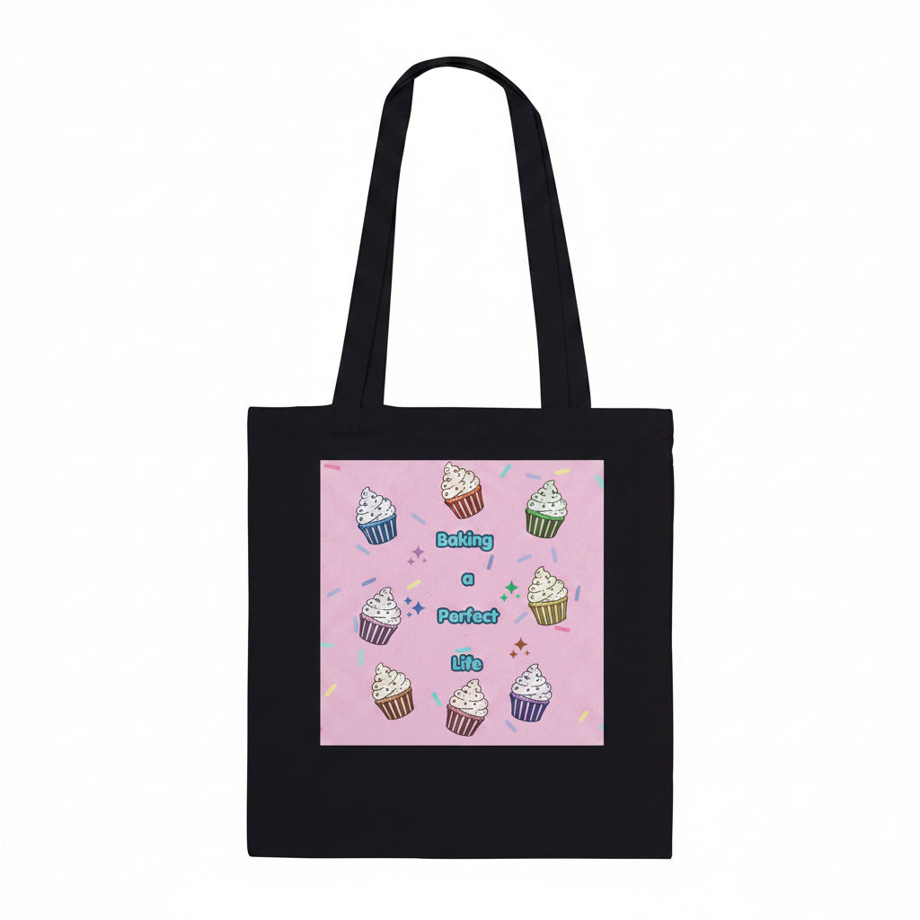 A black canvas tote bag with two shoulder straps, featuring a square pink graphic print on the front. The print displays a cute illustration of colorful cupcakes arranged in a circle around the text 'Baking a Perfect Life' in blue lettering.