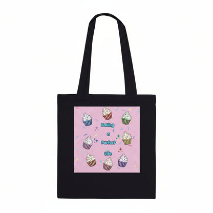 A black canvas tote bag with two shoulder straps, featuring a square pink graphic print on the front. The print displays a cute illustration of colorful cupcakes arranged in a circle around the text 'Baking a Perfect Life' in blue lettering.