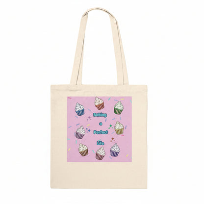 A natural canvas tote bag with a pink square graphic printed on the front, featuring illustrations of cupcakes and the text 'Baking a Perfect life'.