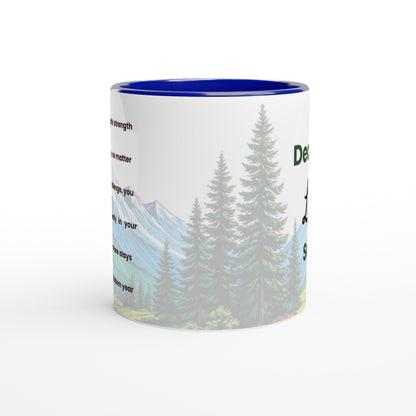 A ceramic mug with a white exterior featuring a scenic graphic of pine trees and mountains, a blue interior, and partial black text printed on the side.