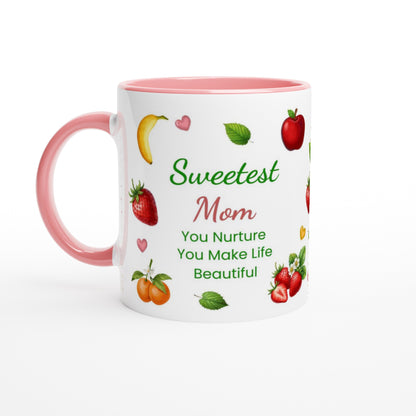A white ceramic mug with a pink handle and pink interior, printed with the phrase 'Sweetest Mom You Nurture You Make Life Beautiful' in green and pink lettering, surrounded by illustrations of fruits and hearts.