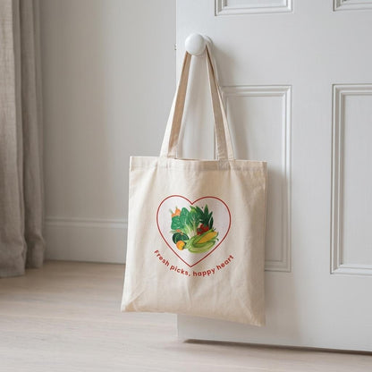 A natural canvas tote bag with a heart graphic of fresh vegetables and the text 'Fresh picks, happy heart' hanging from a white door knob in a bright room.