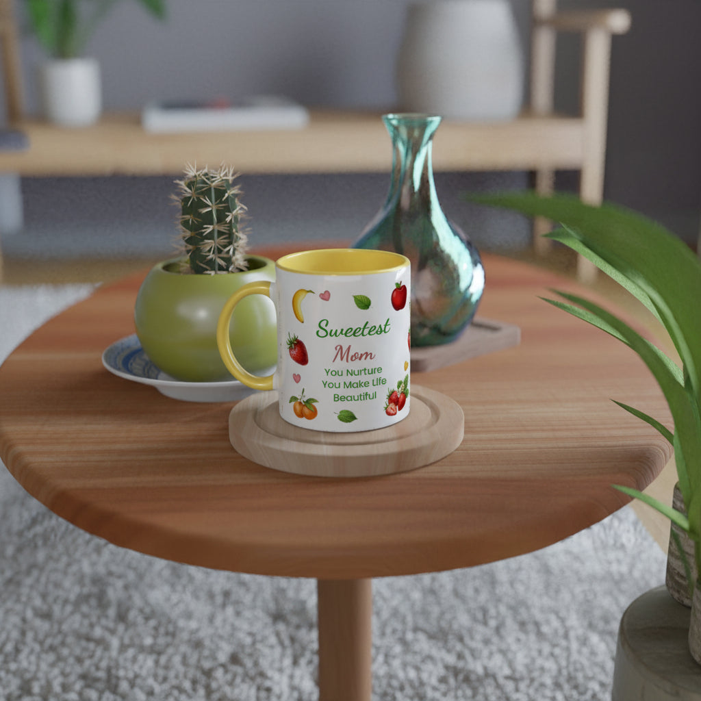 A white ceramic mug with a yellow interior and handle, printed with the phrase 'Sweetest Mom You Nurture You Make Life Beautiful' alongside colorful fruit illustrations, displayed on a round wooden coaster on a wooden table in a cozy, softly blurred living room setting.