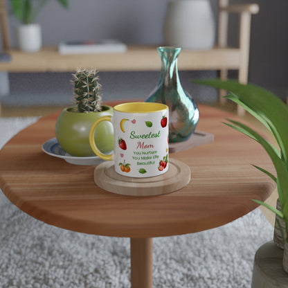 A white ceramic mug with a yellow interior and handle, printed with the phrase 'Sweetest Mom You Nurture You Make Life Beautiful' alongside colorful fruit illustrations, displayed on a round wooden coaster on a wooden table in a cozy, softly blurred living room setting.