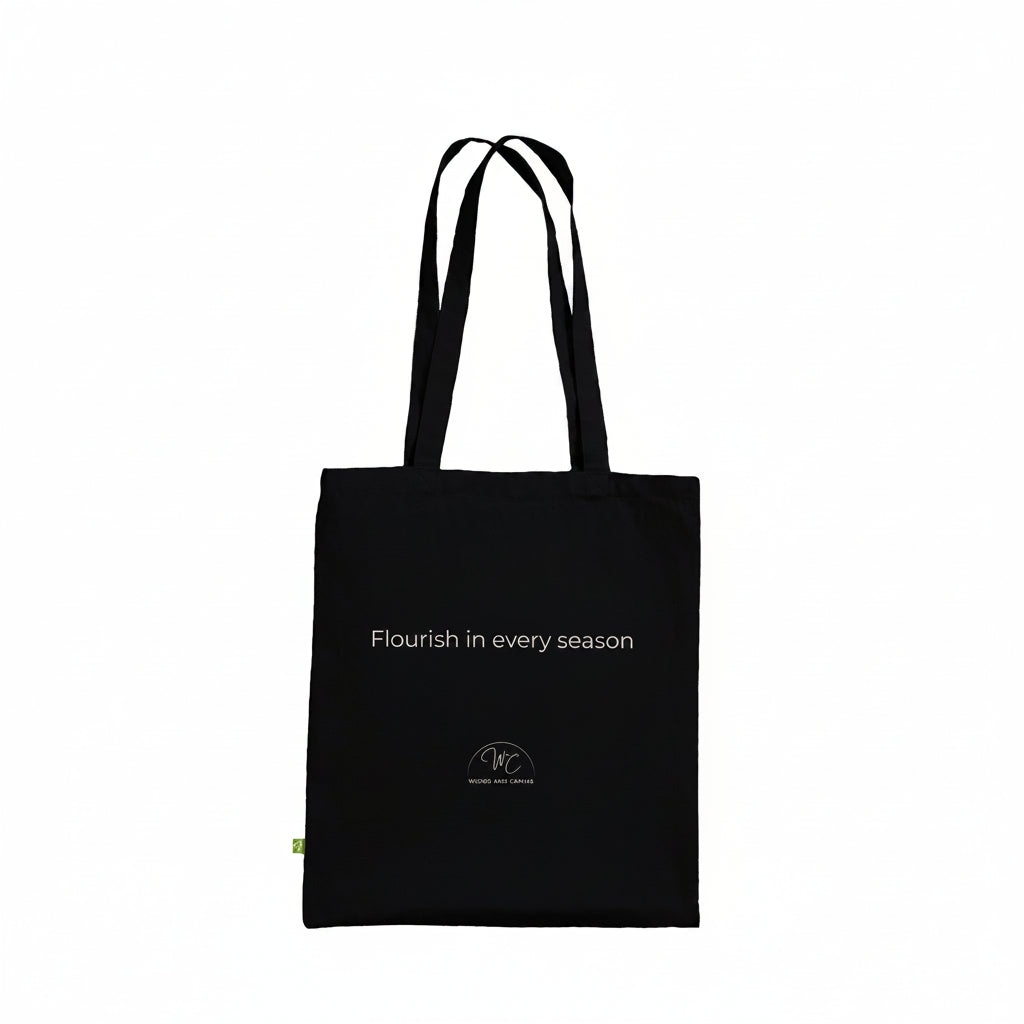 A black canvas tote bag with black shoulder straps and a small gold circular logo on the lower front, shown against a plain white background.