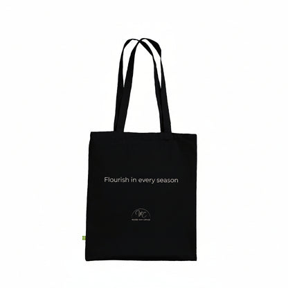 A black canvas tote bag with black shoulder straps and a small gold circular logo on the lower front, shown against a plain white background.