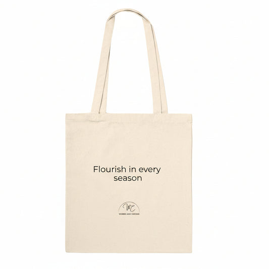 A natural canvas tote bag with black printed text reading 'Flourish in every season' and a small circular logo below, shown on a plain white background.