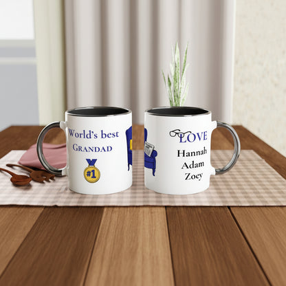 Two personalized white ceramic mugs with black interiors and silver-toned handles, displayed on a wooden table. One mug reads 'World's best GRANDAD' with a gold and blue medal graphic, and the other reads 'LOVE Hannah Adam Zoey' with a blue chair graphic.