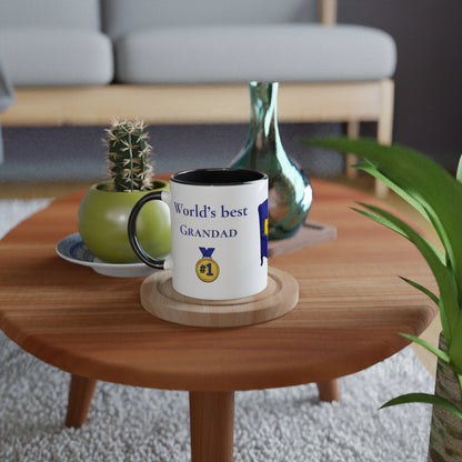 A white ceramic mug with a black handle and rim sits on a round wooden coaster on a wooden coffee table. The mug is printed with the text 'World's best GRANDAD' in blue, along with a gold and blue medal graphic.