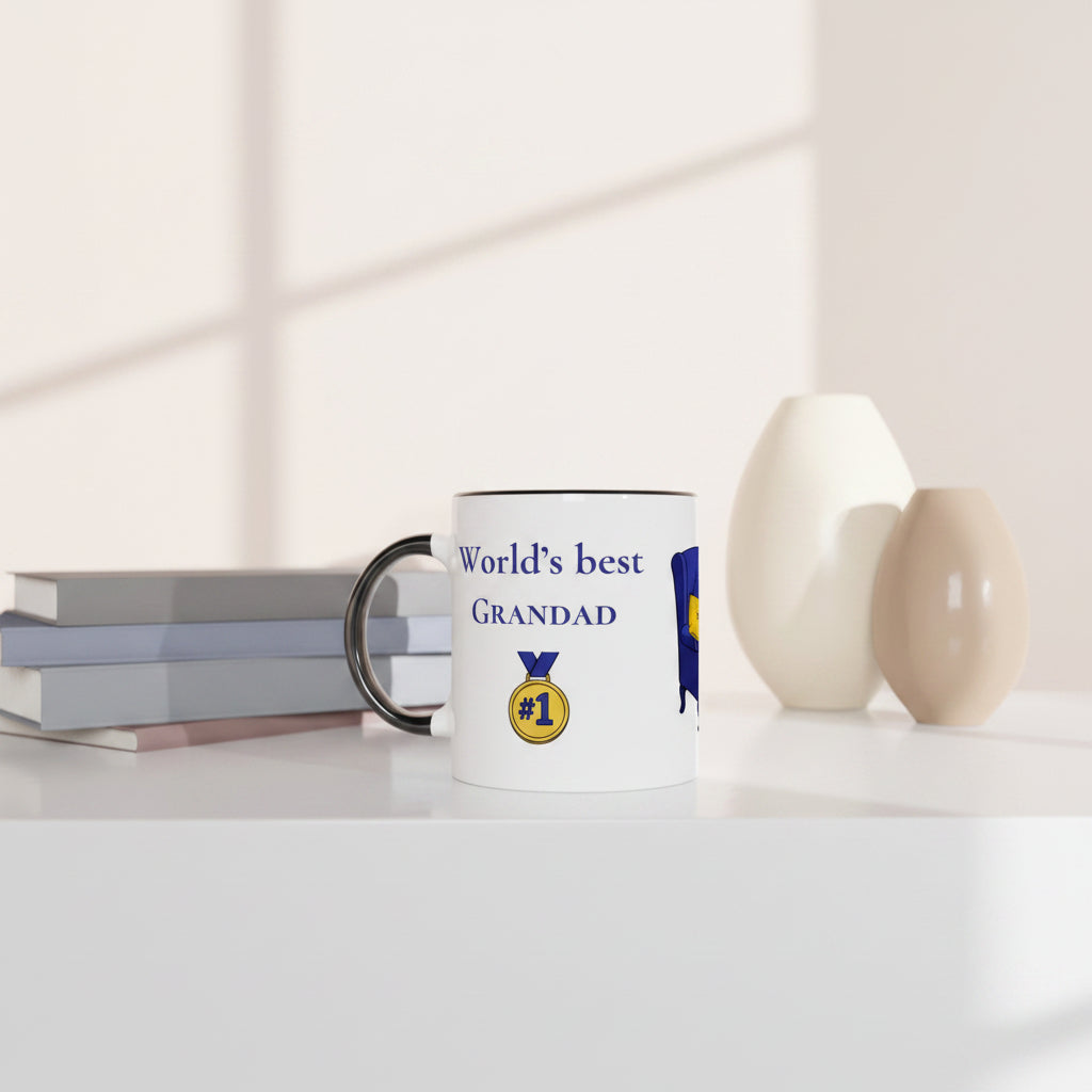 A white ceramic mug with a black handle sits on a white tabletop next to stacked books, with two beige vases in the background. The mug is printed with the text 'World's best GRANDAD' in blue, a gold '#1' medal graphic, and a blue and yellow emblem.