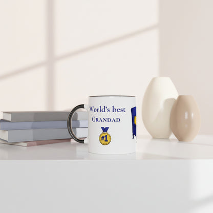A white ceramic mug with a black handle sits on a white tabletop next to stacked books, with two beige vases in the background. The mug is printed with the text 'World's best GRANDAD' in blue, a gold '#1' medal graphic, and a blue and yellow emblem.