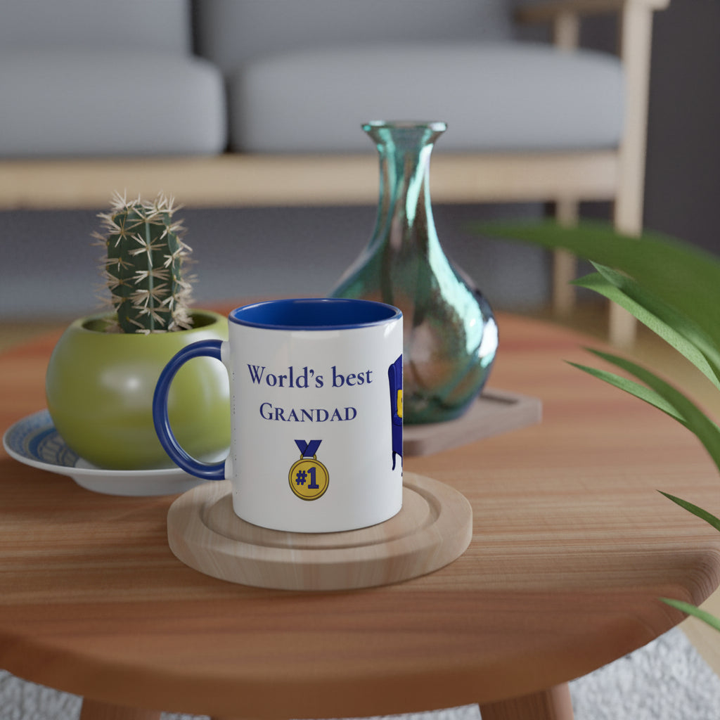 A white ceramic mug with a blue handle and interior sits on a round wooden coaster on a wooden table. The mug is printed with the text 'World's best GRANDAD' and a gold '#1' medal graphic.