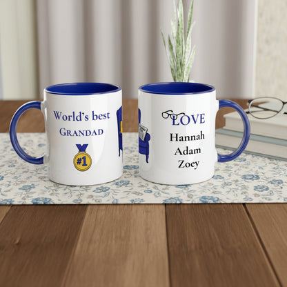 Two personalized white ceramic mugs with blue interiors and handles, displayed side by side on a checkered placemat. The left mug reads 'World's best GRANDAD' with a gold medal graphic, and the right mug reads 'LOVE' above the names 'Hannah', 'Adam', and 'Zoey'.