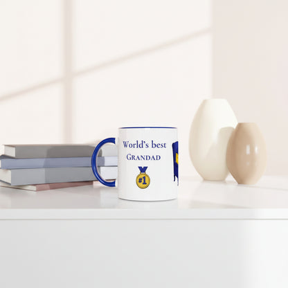 A white ceramic mug with a blue handle and rim, printed with the text 'World's best GRANDAD' and a gold medal graphic, styled on a white shelf next to books and two vases.
