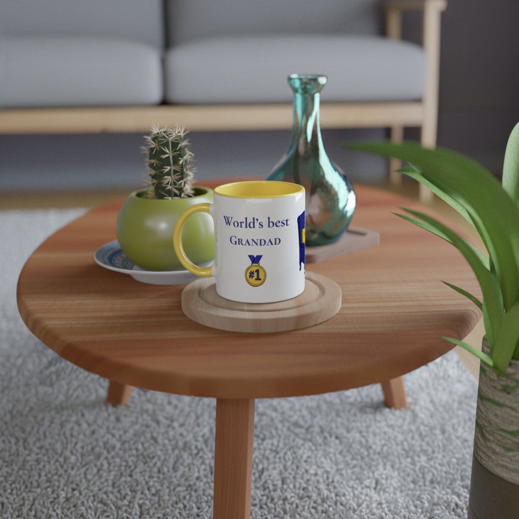 A white ceramic mug with a yellow interior and handle sits on a round wooden coaster on a round wooden coffee table. The mug is printed with the text 'World's best GRANDAD' and a gold medal emblem.