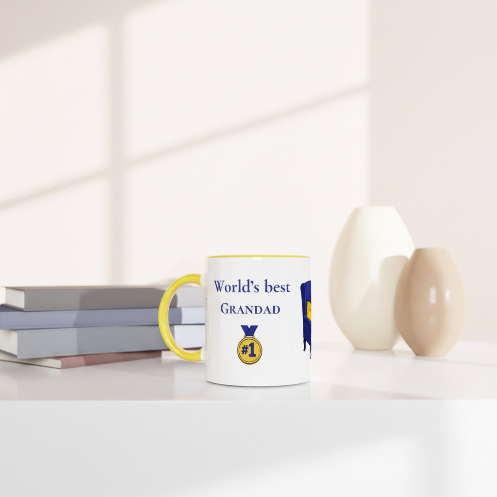 A white ceramic mug with a yellow handle and rim, printed with the text 'World's best GRANDAD' and a gold medal graphic, styled on a white table next to books and two ceramic vases.