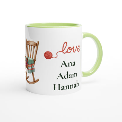 A white ceramic mug with a light green handle and interior, featuring a printed design of a wooden rocking chair with a green blanket, a red ball of yarn, the word 'love' in red script, and the names 'Ana', 'Adam', and 'Hannah' 