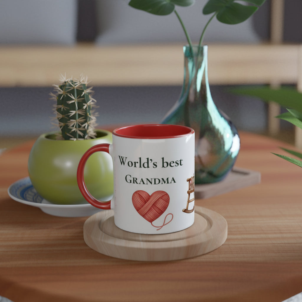 A white ceramic mug with a red interior and handle sits on a wooden coaster on a wooden table. The mug is printed with the text 'World's best GRANDMA' and a red heart made of yarn.