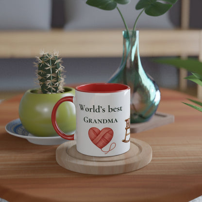 A white ceramic mug with a red interior and handle sits on a wooden coaster on a wooden table. The mug is printed with the text 'World's best GRANDMA' and a red heart made of yarn.