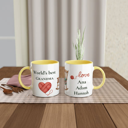 Two personalized ceramic mugs with yellow interiors and handles, displayed on a wooden table with a placemat and small plant. One mug reads 'World's best GRANDMA' with a red heart of yarn, and the other reads 'love Ana Adam Hannah