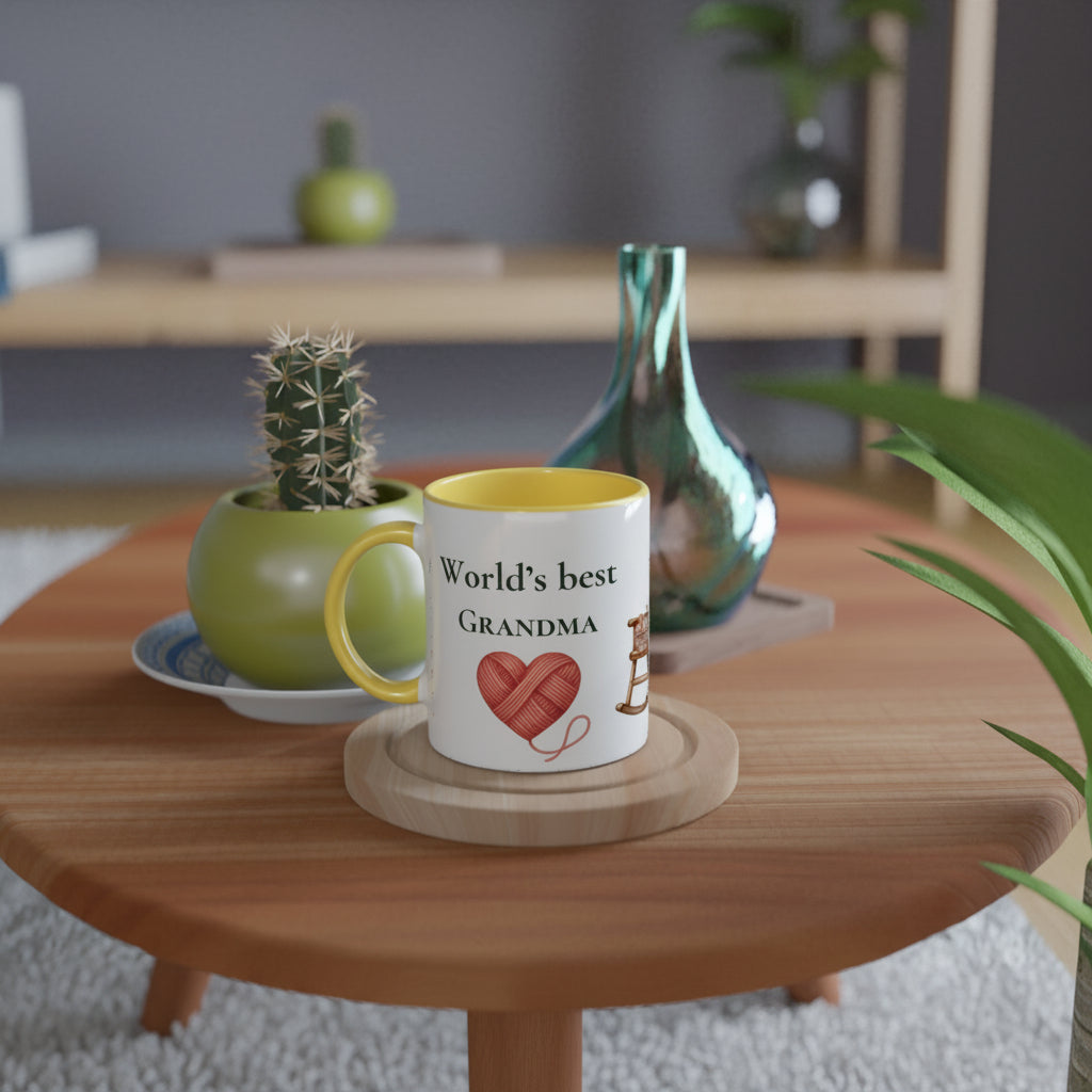 A white ceramic mug with a yellow handle and interior sits on a round wooden coaster on a light wood table. The mug is printed with the text 'World's best GRANDMA' and a red heart made of yarn.