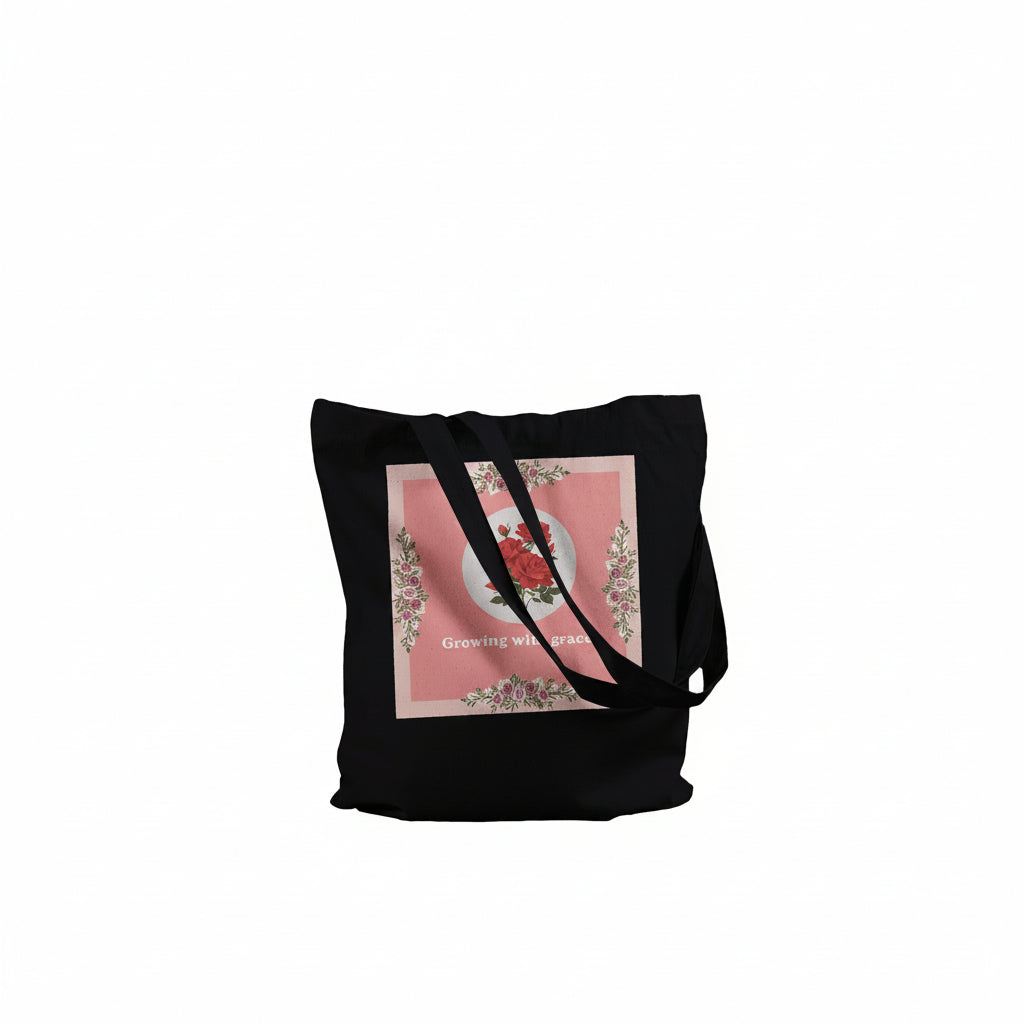 A black canvas tote bag with long shoulder straps, featuring a square pink graphic print on the front with a floral wreath design and the text 'Growing with grace'. 