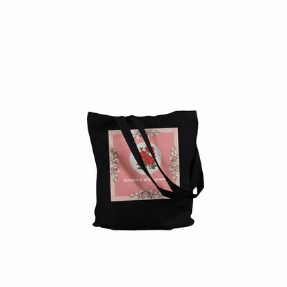 A black canvas tote bag with long shoulder straps, featuring a square pink graphic print on the front with a floral wreath design and the text 'Growing with grace'. 
