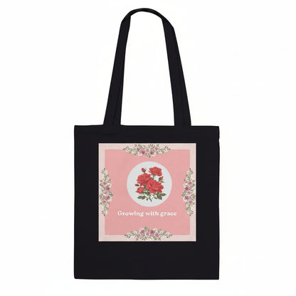 A black canvas tote bag with two shoulder straps, featuring a square pink floral graphic on the front with the text 'Growing with grace'.