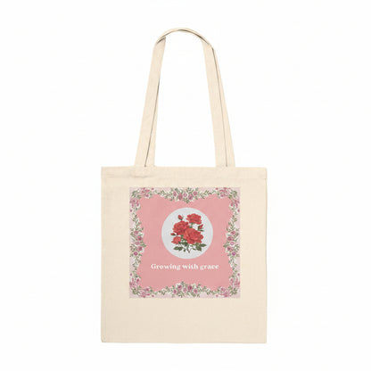 A natural canvas tote bag with a pink floral graphic on the front featuring red roses and the text 'Growing with grace'.