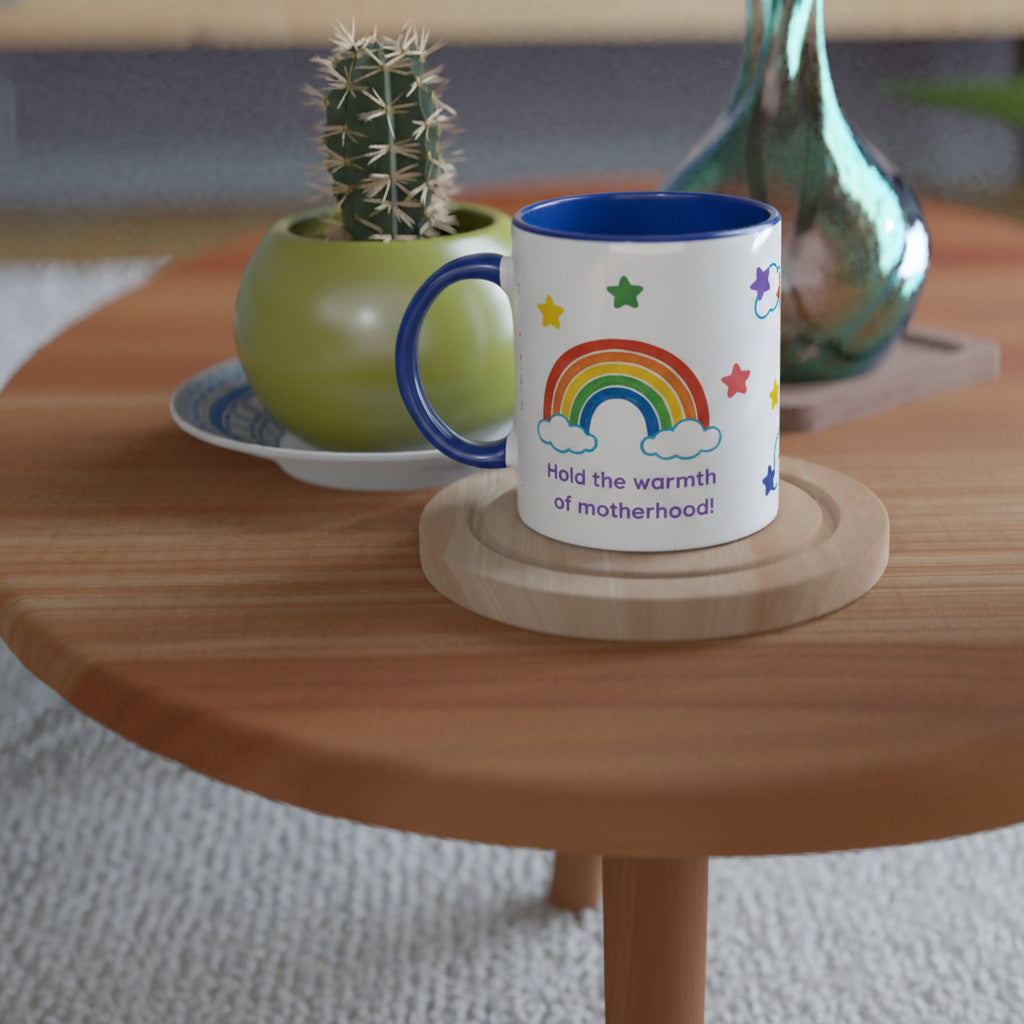 A white ceramic mug with a blue handle and interior, printed with a colorful rainbow, stars, and the phrase 'Hold the warmth of motherhood!', sits on a round wooden coaster on a wooden table. A small potted cactus and other decor are blurred in the background.