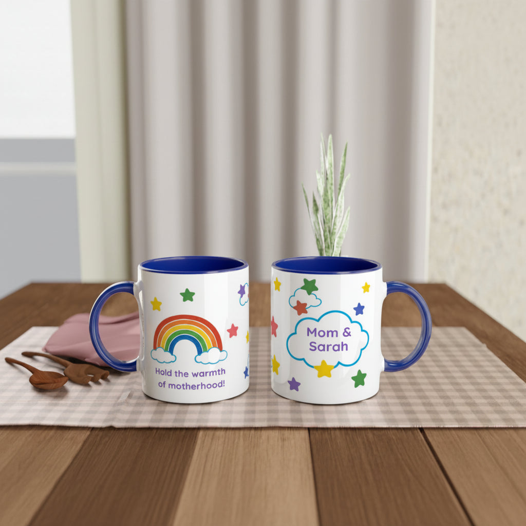 Two white ceramic mugs with blue interiors and handles sit on a wooden table. The left mug has a rainbow, clouds, and colorful stars with the text 'Hold the warmth of motherhood!'.